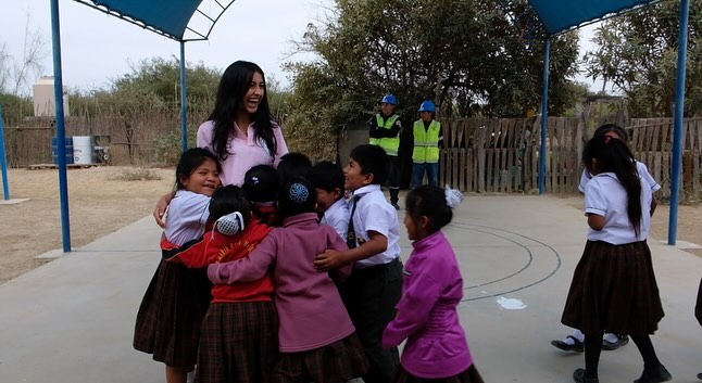 A group of young children in school uniforms hug a smiling woman, Sydney Assil, under a blue canopy outdoors, while three adults in safety vests and helmets from Sweet Impact stand in the background near a fence.