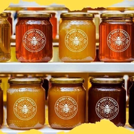 Six jars of honey with different shades of golden and amber color, arranged on two shelves. The jars have gold lids and labels that read My Honey Project with a bee graphic in the center.