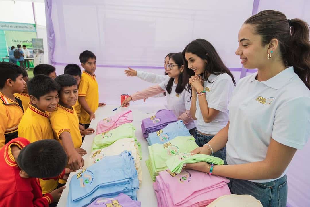 Two women stand behind a table covered with colorful folded T-shirts, smiling and talking to a group of children in yellow uniforms, who are looking at the shirts and reaching out to pick one.