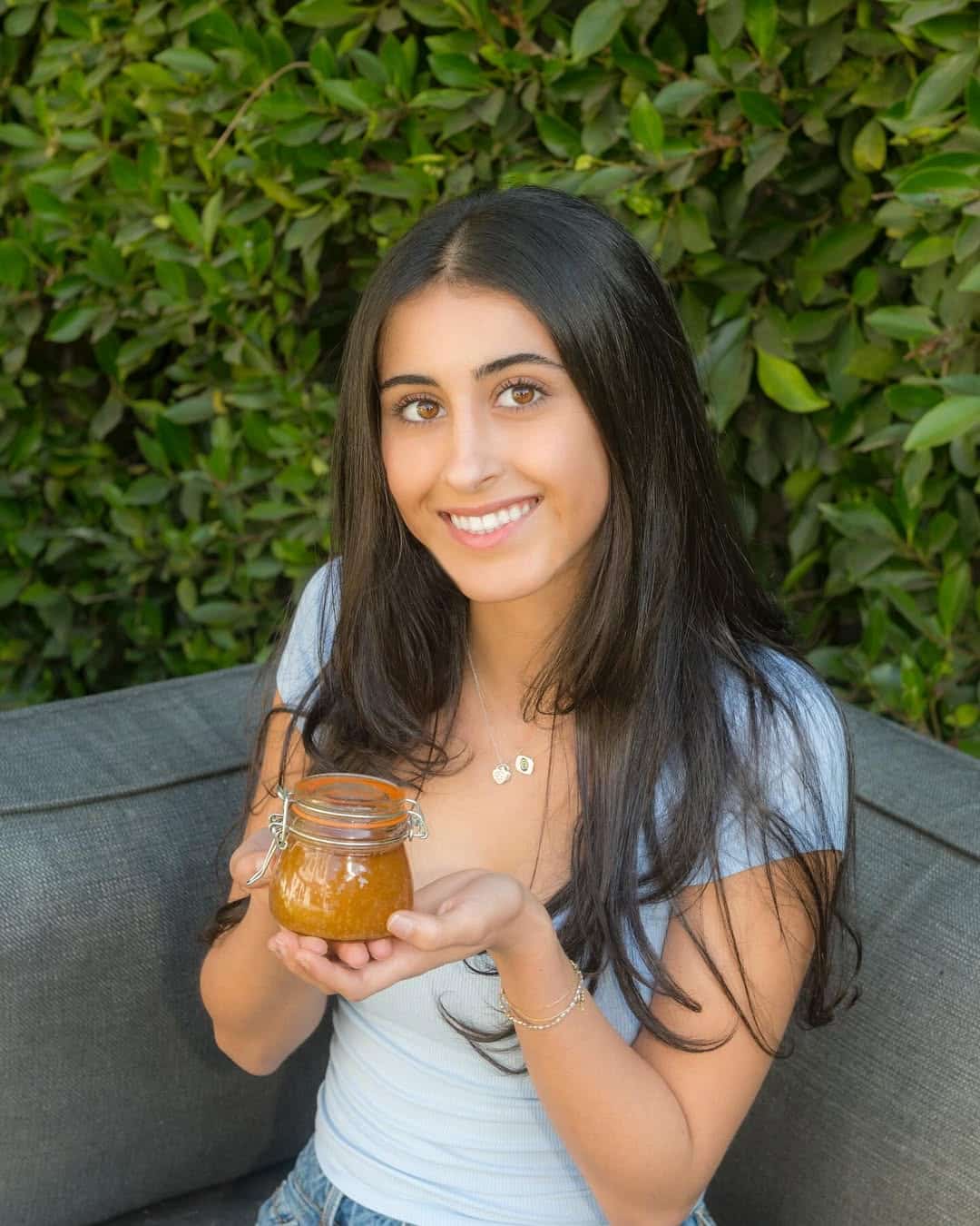 A young woman with long dark hair, wearing a light blue top, sits on a gray couch outdoors and smiles while holding a small glass jar filled with brownish-orange contents. Green leaves fill the background.