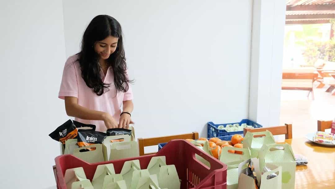 A woman with long dark hair, wearing a light pink shirt, smiles as she packs food items into green boxes on a table. There are oranges and other supplies around her in trays and baskets.