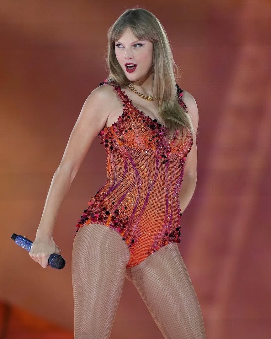 A woman with long blonde hair performs on stage wearing a sparkling orange and red bodysuit, holding a microphone in her right hand, with a dramatic expression and an orange-lit background.