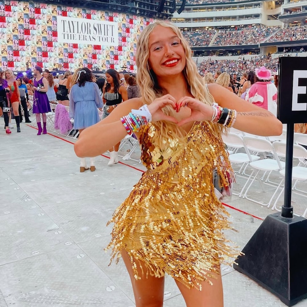 A woman in a gold sequin dress smiles and makes a heart shape with her hands at a stadium concert. The stage behind her displays Taylor Swift The Eras Tour, while fans in colorful DIY costumes inspired by Taylor Swift fill the background.