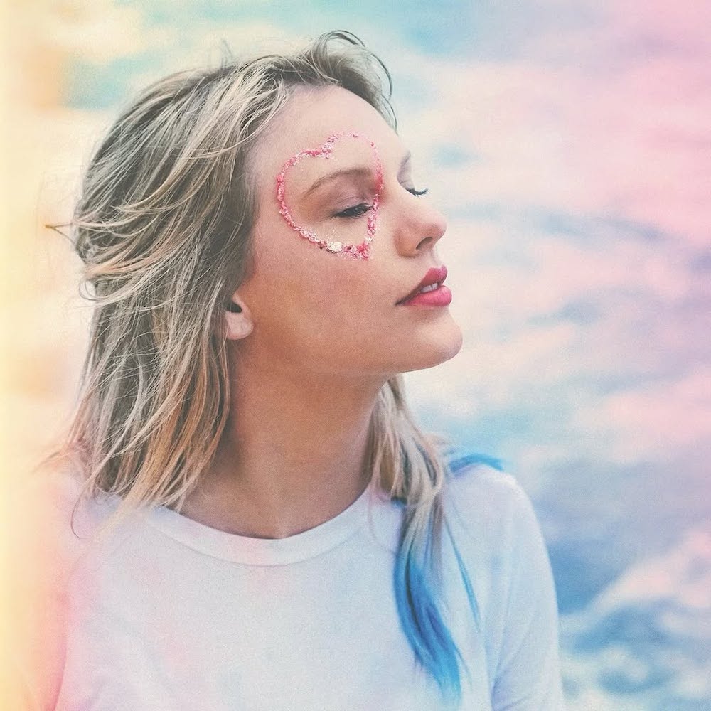 A woman with long blonde hair and closed eyes stands peacefully, evoking a dreamy Taylor Swift album vibe. She has a pink heart design around her right eye, wears a white shirt, and the pastel background is softly blurred.