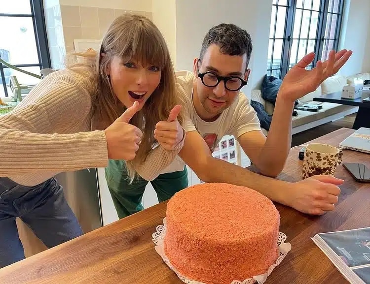 A woman with long hair gives two thumbs up next to a man with glasses who is shrugging. Both are sitting at a table with a pink cake—possibly inspired by Taylor Swift’s recipe dupes—in a bright room with large windows.
