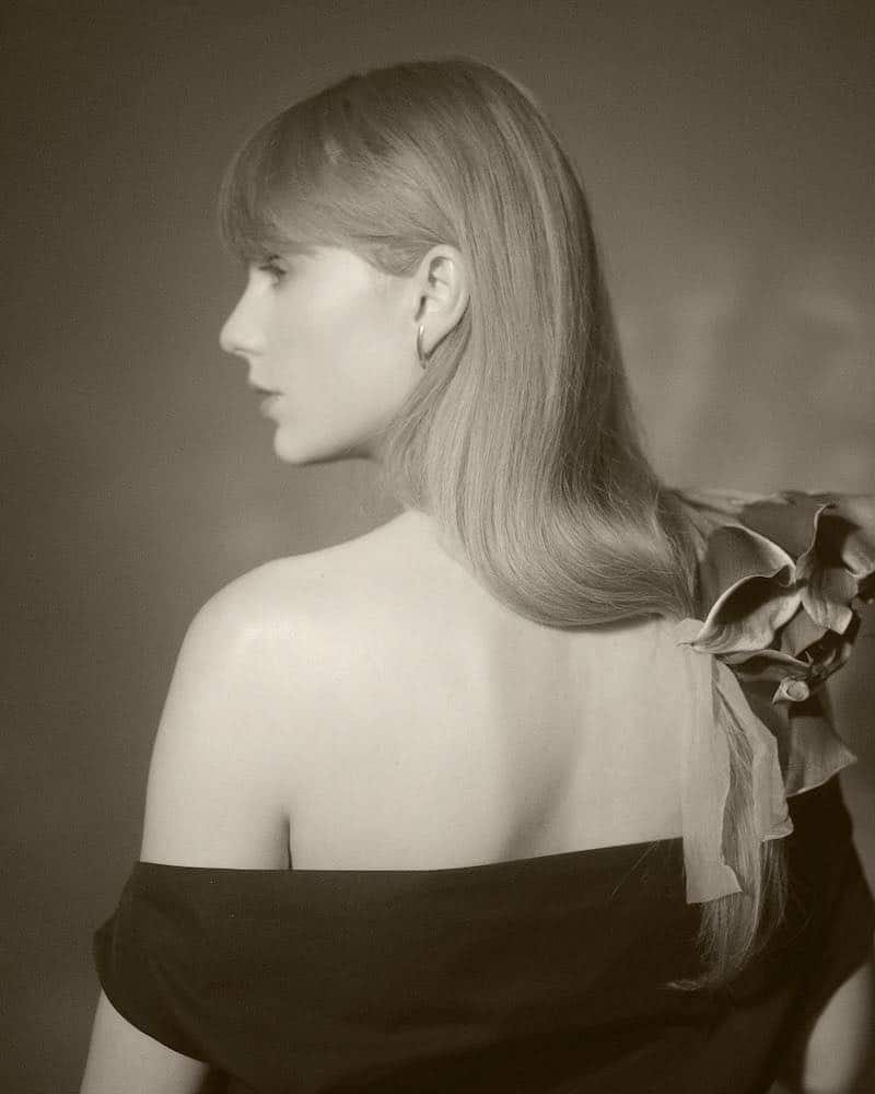 A woman with long, straight hair looks to the side, evoking the mood of a Taylor Swift album. She wears an off-the-shoulder black top and a large fabric flower. The black and white portrait features soft lighting and a blurred background.