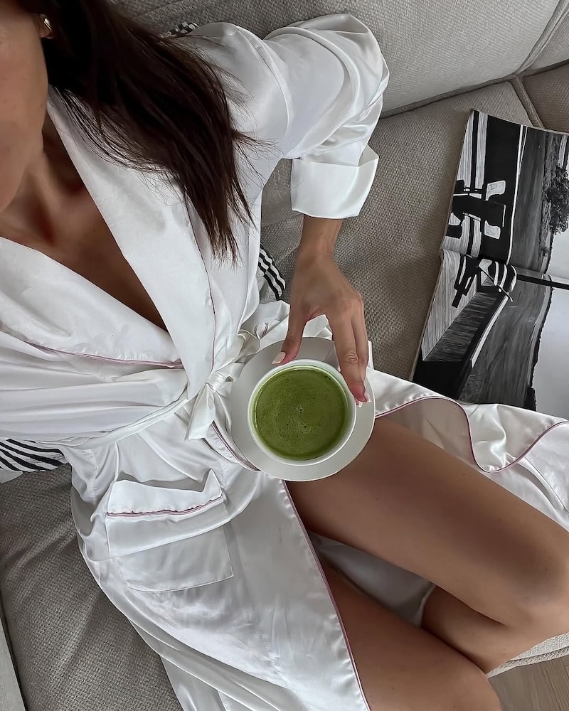 A woman in a white satin robe sits on a couch holding a cup of green matcha latte, enjoying an at-home spa day. A black and white photo book rests beside her. Only her lower face and body are visible.
