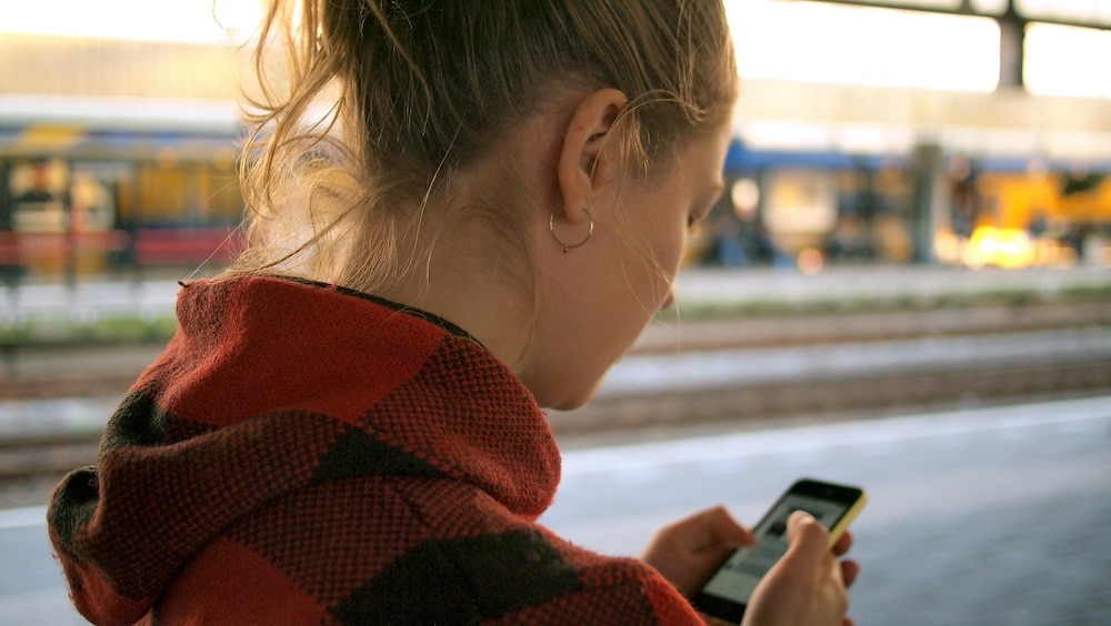 A woman with blond hair in a ponytail, wearing a red plaid jacket, looks at her smartphone—perhaps reading about the social media ban Australia debates—while standing on a train platform with tracks and a blurred train in the background.