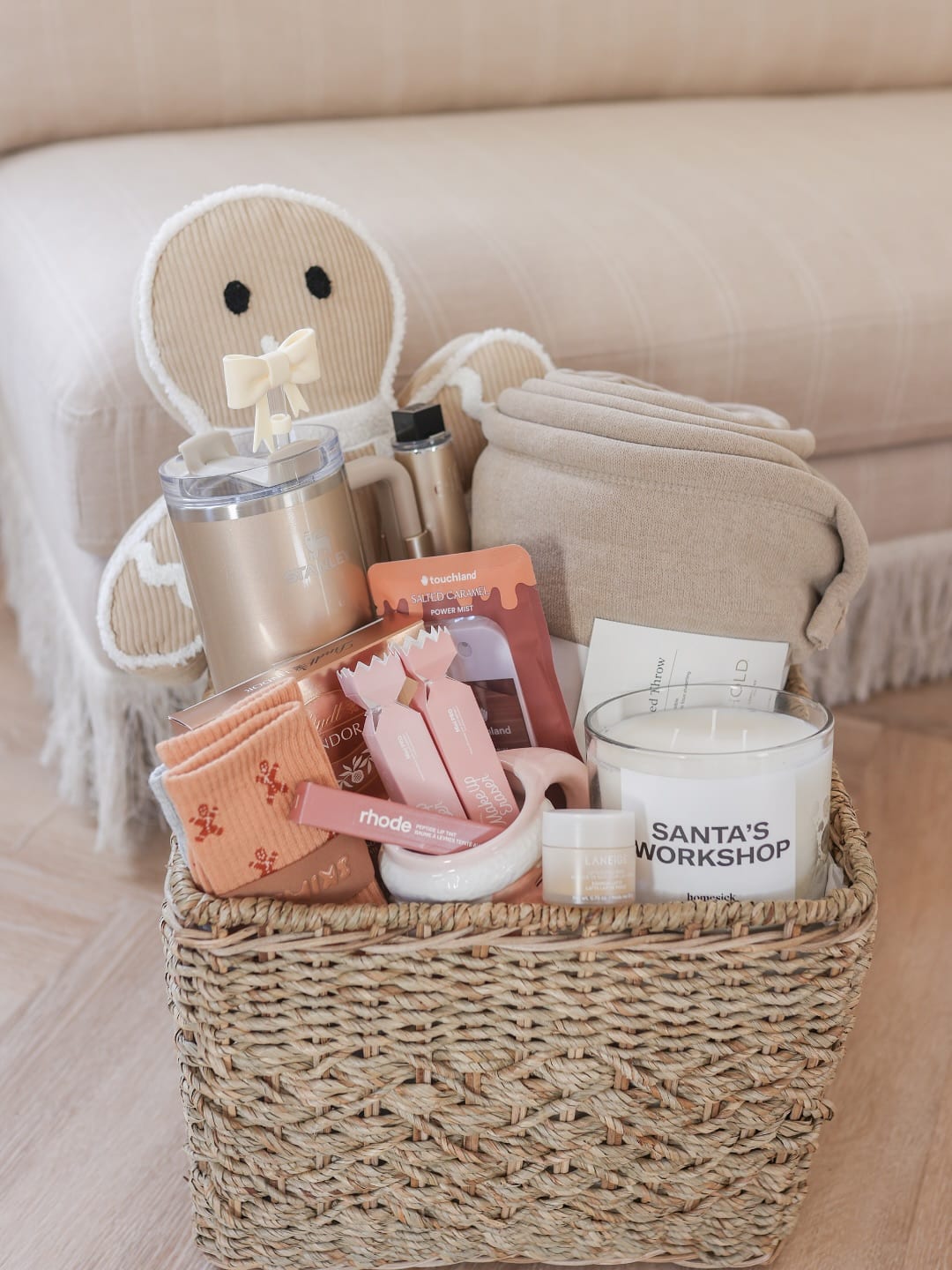 A woven basket filled with various gift items, including a plush doll, a tumbler, cozy socks, skincare products, a blanket, a candle labeled “Santa’s Workshop,” and other small self-care essentials.