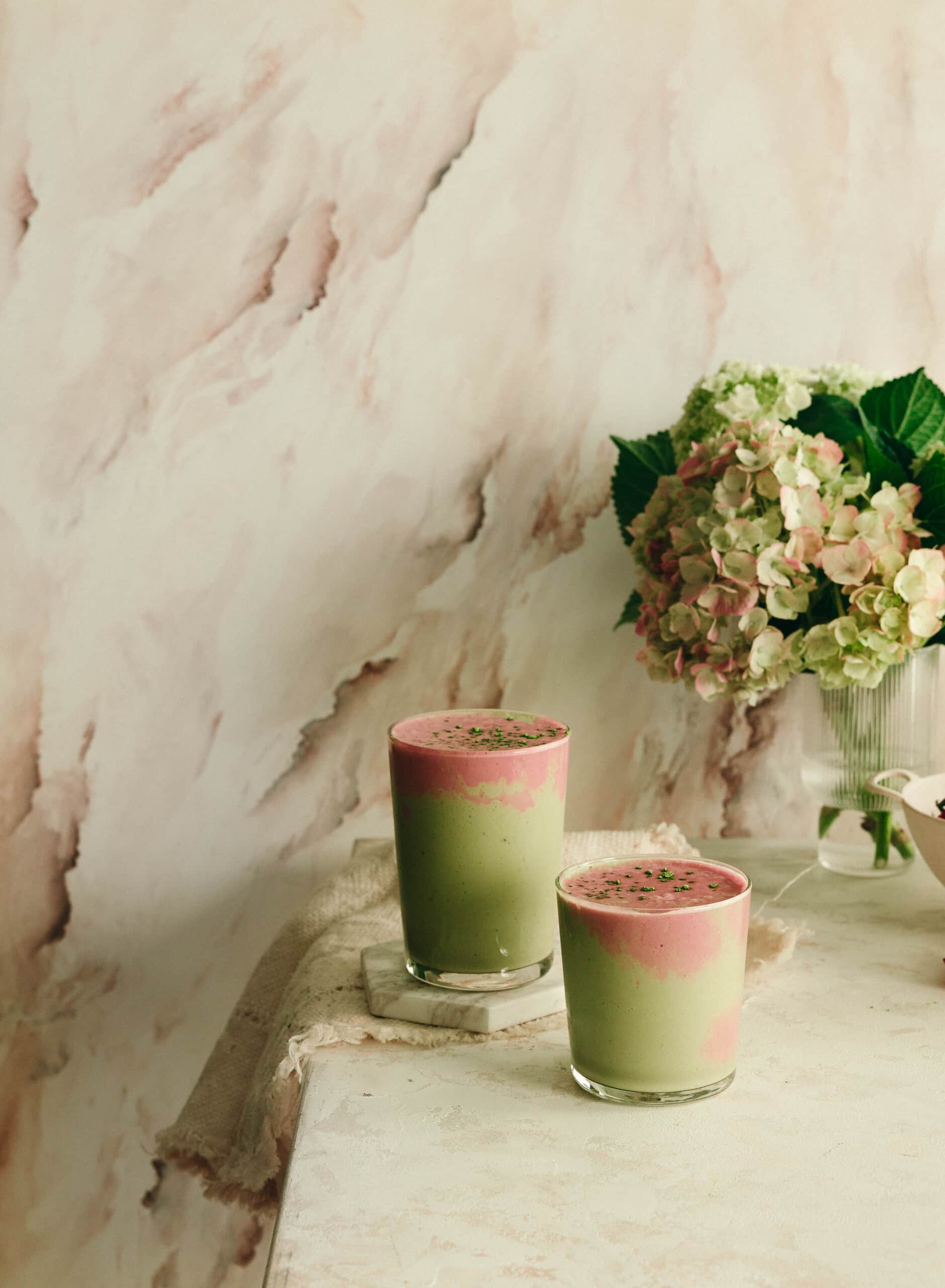 Two glasses filled with layered green and pink smoothies—perfect healthy snacks—sit on a light-colored table next to a vase of hydrangeas, with a marble-patterned wall in the background.