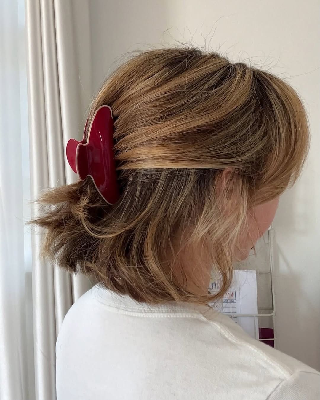 A person with short, wavy, light brown hair is wearing a large red claw clip as a hair accessory at the back of their head. They are facing away from the camera, dressed in a white shirt and standing near white curtains.