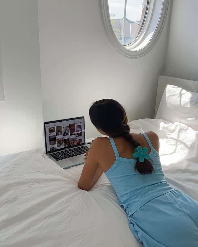 A person in a light blue outfit with a blue flower hair clip lies on a white bed, using a laptop to research how to start therapy. Sunlight streams through a round window above, brightening the minimalist white room.