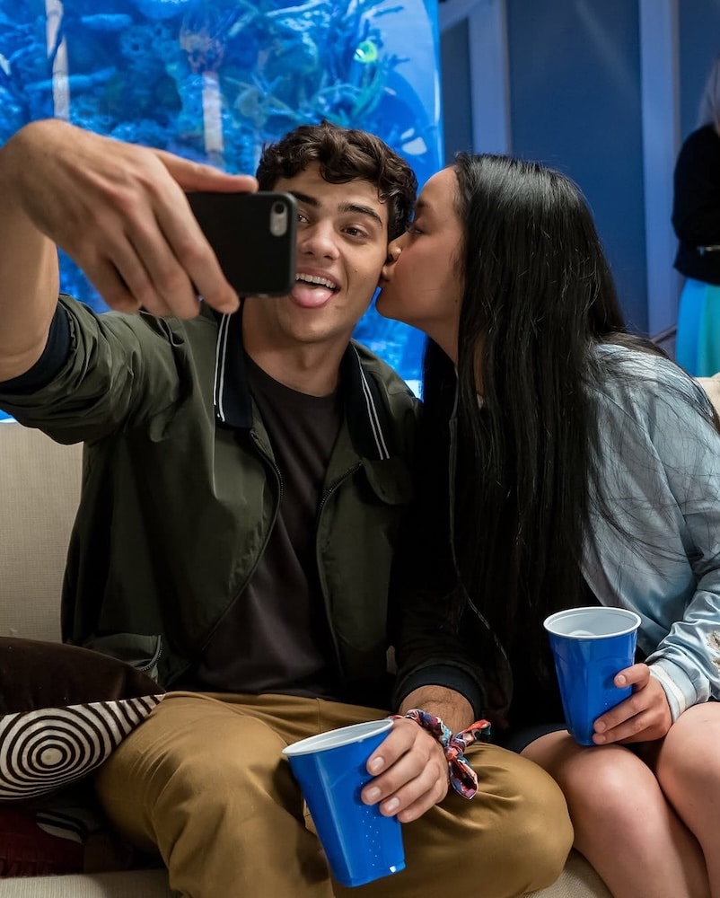 A young man and woman sit on a couch holding blue cups; the woman kisses the man's cheek while he takes a selfie. Both appear happy and playful, like a scene from romantic comedies, with a large aquarium in the background.