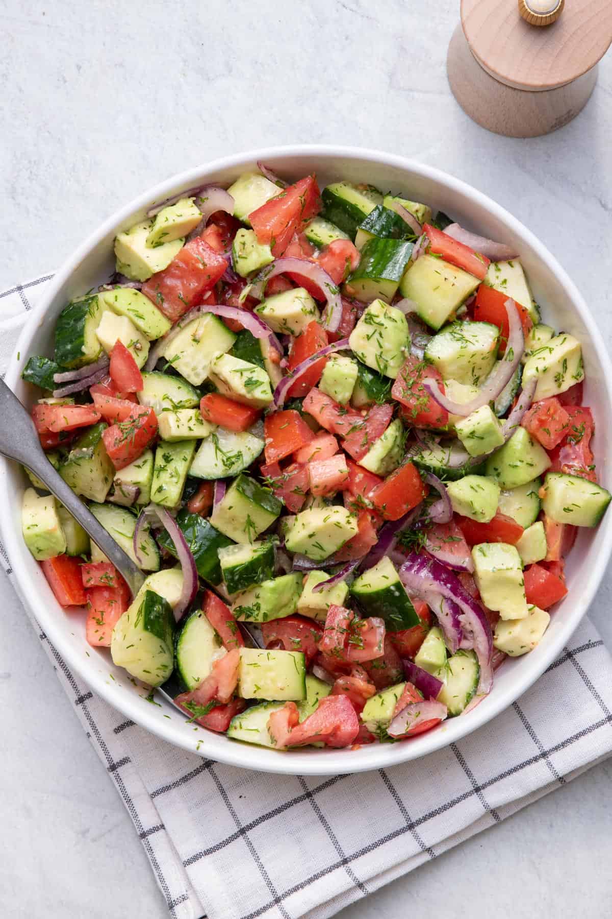 A bowl of fresh salad with chopped cucumbers, tomatoes, red onions, and avocado, garnished with herbs. Perfect for healthy snacks or after school snacks, a serving spoon rests in the bowl on a checkered cloth on a light surface.