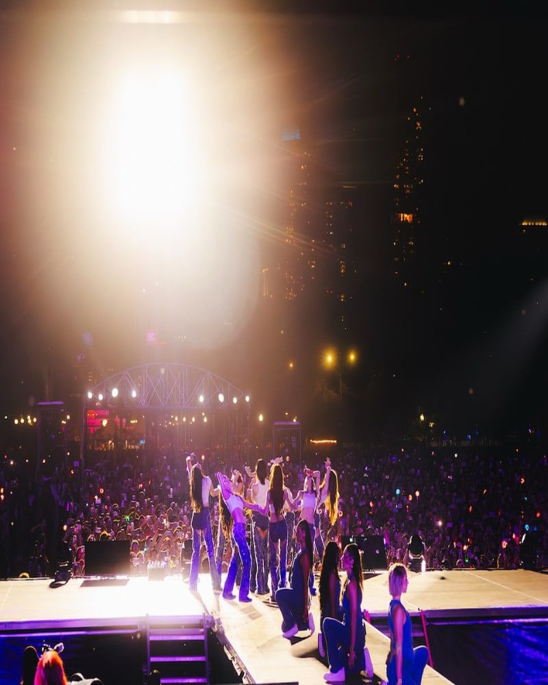 A music group performs on an outdoor stage at night, facing a large cheering crowd under bright stage lights with a city skyline visible in the background.