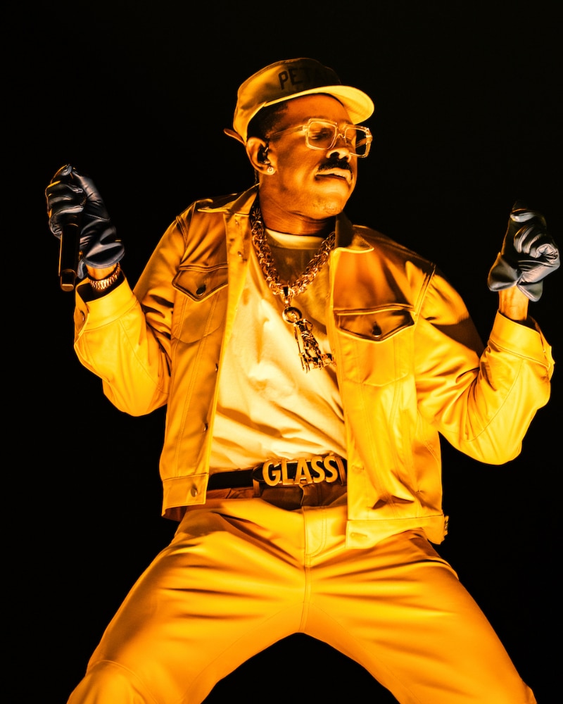 A man wearing yellow pants, a yellow jacket, hat, sunglasses, and gloves performs on stage holding a microphone. He is illuminated by dramatic yellow lighting and wears a thick chain necklace and a belt reading “GLASSY.”.