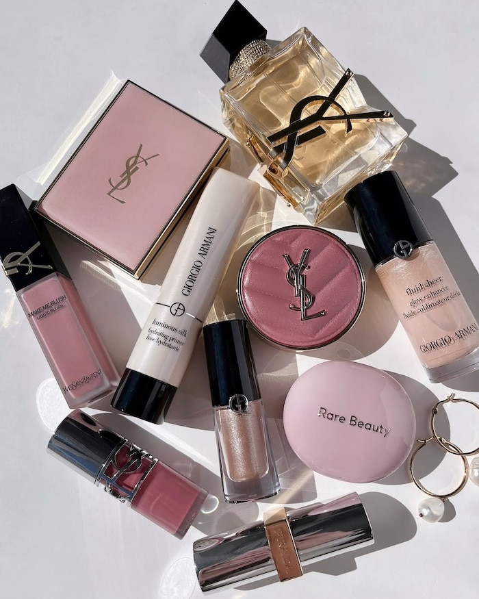 A flat lay of various luxury makeup products from brands like YSL, Giorgio Armani, and Rare Beauty, including lipsticks, foundation, blush, highlighter, perfume, powder compact, and gold hoop earrings on a white surface.