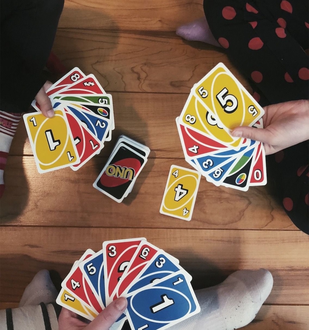 Three people sit on a wooden floor playing UNO, each holding colorful cards. The UNO pile sits center stage, and someone sports red polka-dot pajama pants—perfect for game night fun or as last-minute gift ideas for friends who love games.