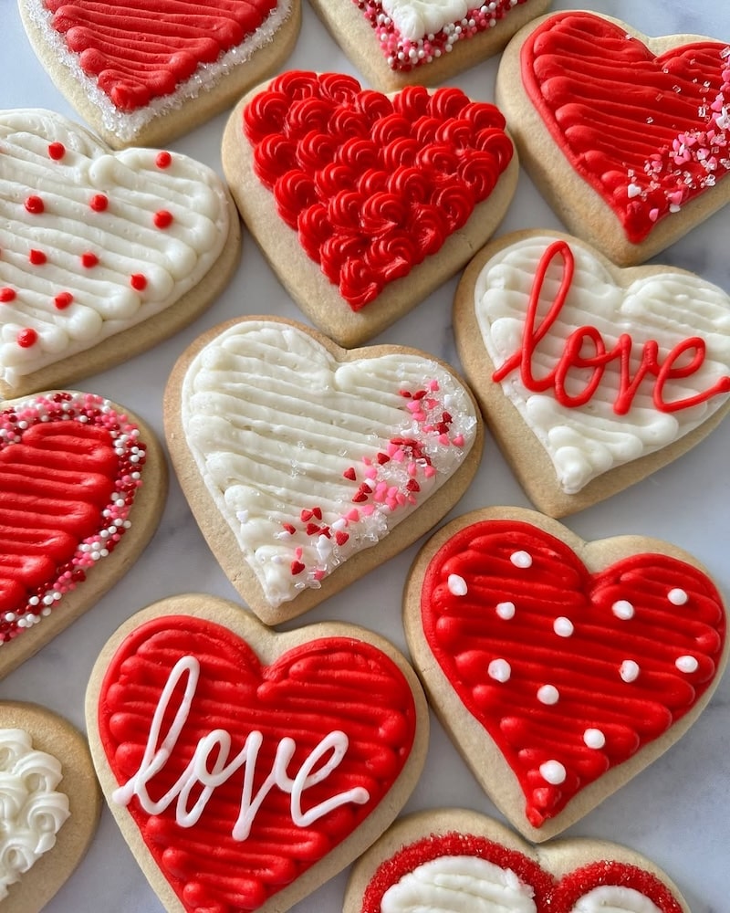 Heart-shaped sugar cookies decorated with red and white icing, sprinkles, and the word love—perfect treats for a Galentine's Day party. Some cookies feature polka dots or swirl patterns, while others are topped with tiny heart-shaped sprinkles.