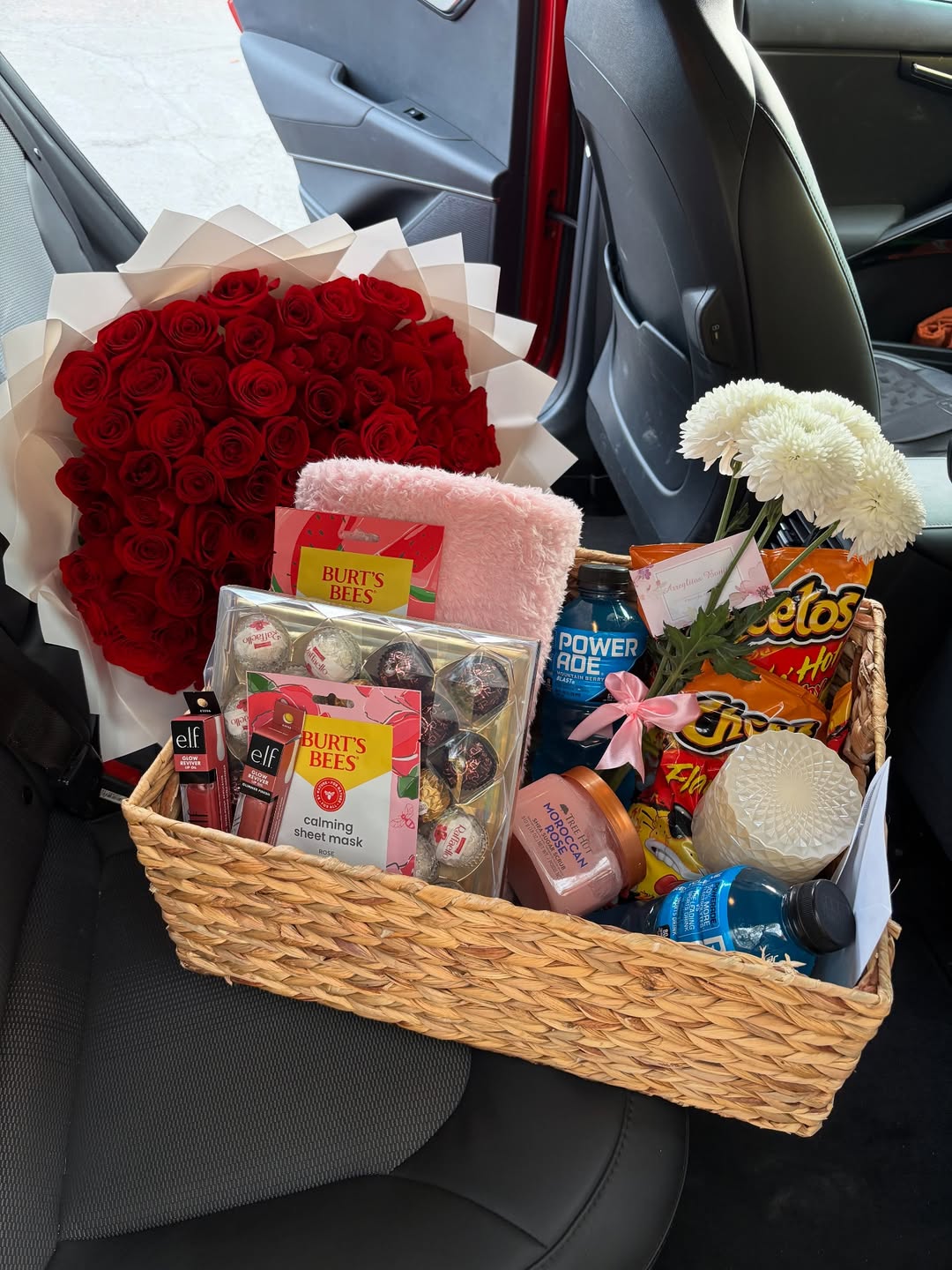 A V-day basket sits on the car seat with snacks, skincare products, a notebook, chocolates, and drinks—all thoughtfully gathered on a budget. Beside it are a bouquet of red roses and a smaller bunch of white flowers.