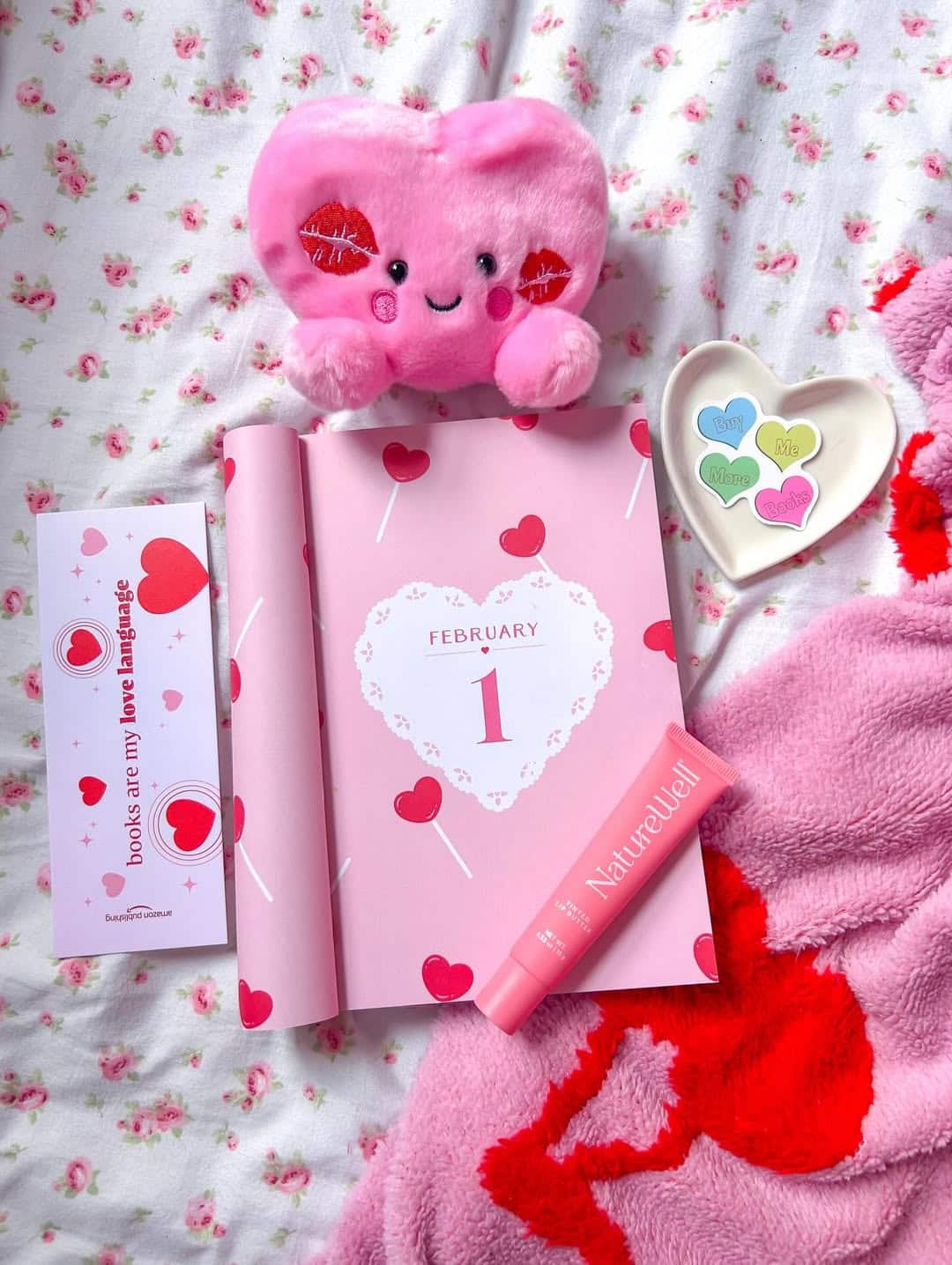 A pink February calendar opened to the 1st, surrounded by a heart plush toy, V-Day bookmark, heart-shaped candies in a dish, a pink tube of cream, and cozy blanket—all set on a floral bedsheet. Looks like February is booked for love!.
