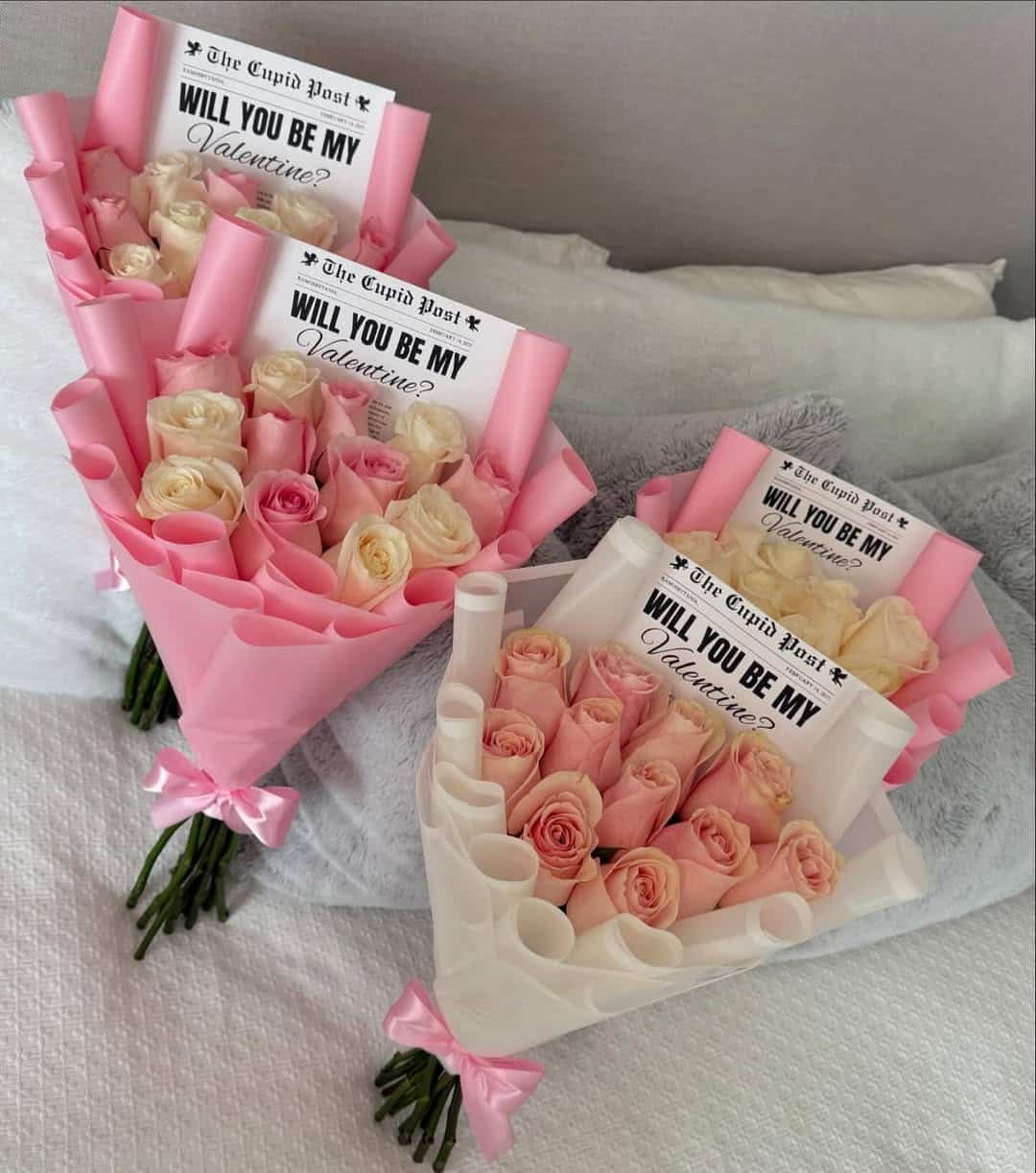 Three bouquets of pink and cream roses, each wrapped in pastel paper with a pink ribbon and a card reading “WILL YOU BE MY Valentine?”—a perfect Valentine's Day budget gift displayed on a soft surface with grey pillows in the background.