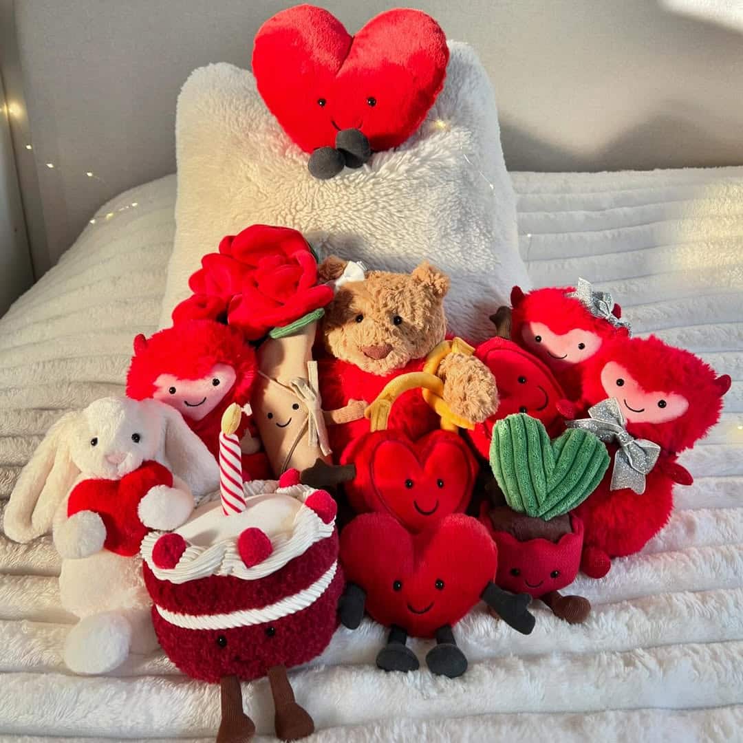 A group of cute plush toys, perfect for a V-day basket on any budget, includes red hearts, a teddy bear, a rabbit, cake slice, rose, and heart-shaped plants—all with smiling faces—arranged on a white, fluffy bed.