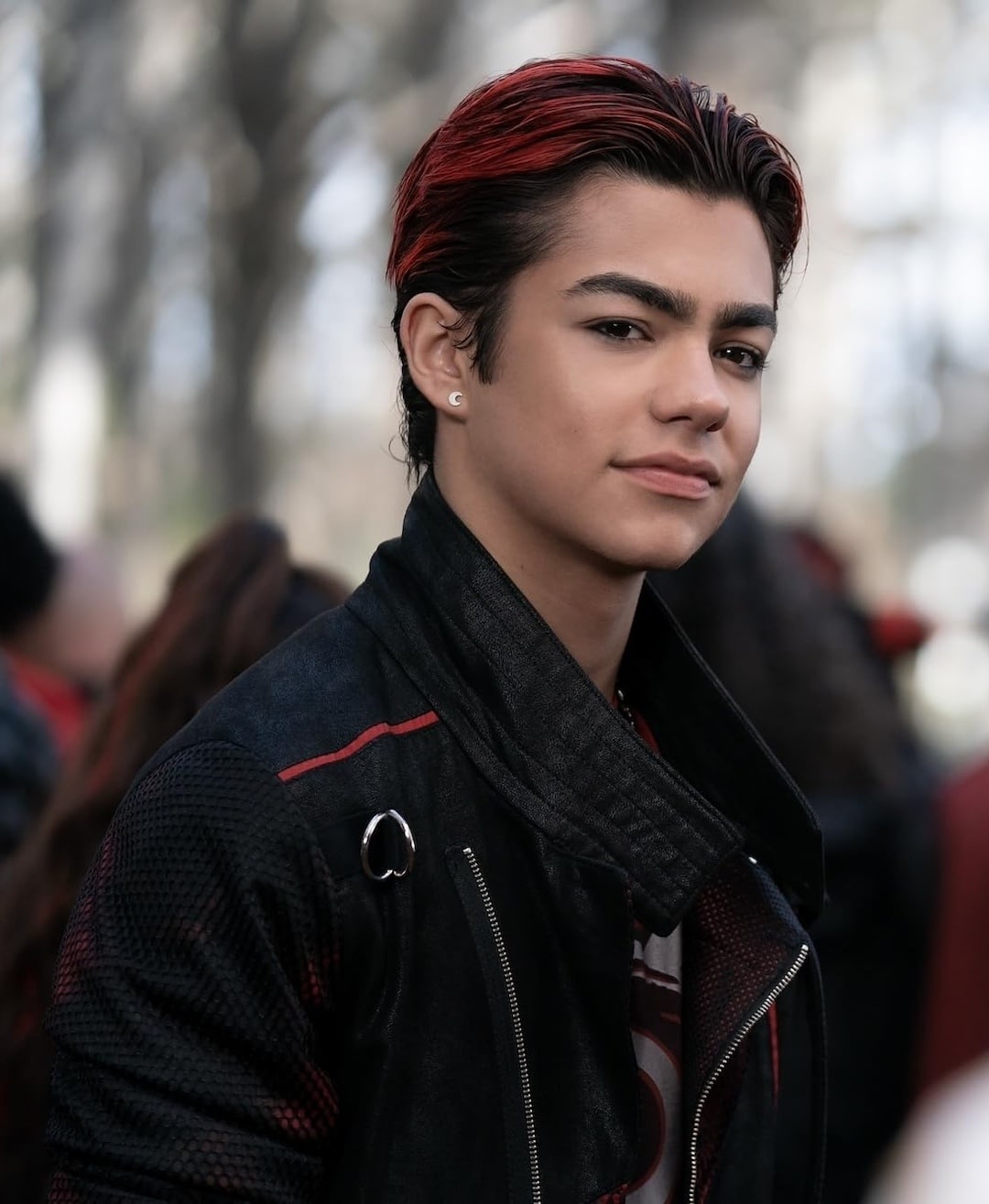 A young man with slicked-back, short dark hair with red highlights, wearing a black jacket with a silver pin and a white earring, stands outdoors with a confident expression—reminiscent of Conrad Fisher on Valentine's Day. The background is blurred with trees and people.