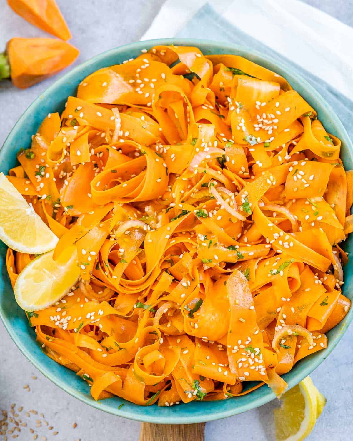 A bowl of carrot ribbon salad garnished with sesame seeds, fresh herbs, and lemon wedges, served in a blue bowl on a light surface—a perfect choice for healthy snacks or after school snacks.