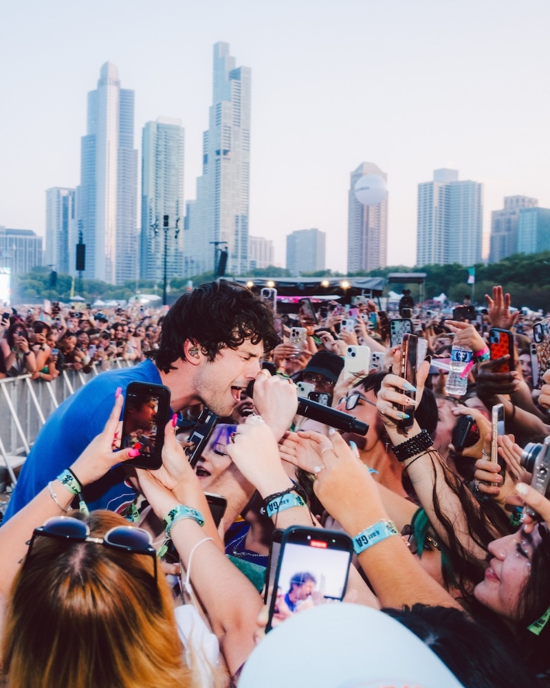 A singer performs passionately in front of a large, excited crowd at an outdoor concert. Fans hold up their phones to take photos, with city skyscrapers visible in the background.