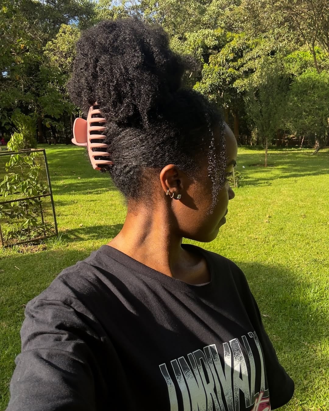 A person with natural curly hair in a loose bun held by a large pink claw clip stands outdoors in sunlight, wearing a black graphic t-shirt and small earrings, with green grass and trees in the background.