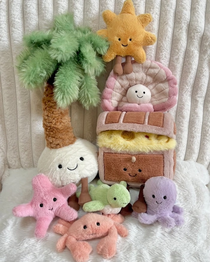 A group of plush toys shaped like sea creatures, a palm tree, a sun, and a treasure chest are arranged together on a white fuzzy surface, all featuring smiling faces and pastel colors.