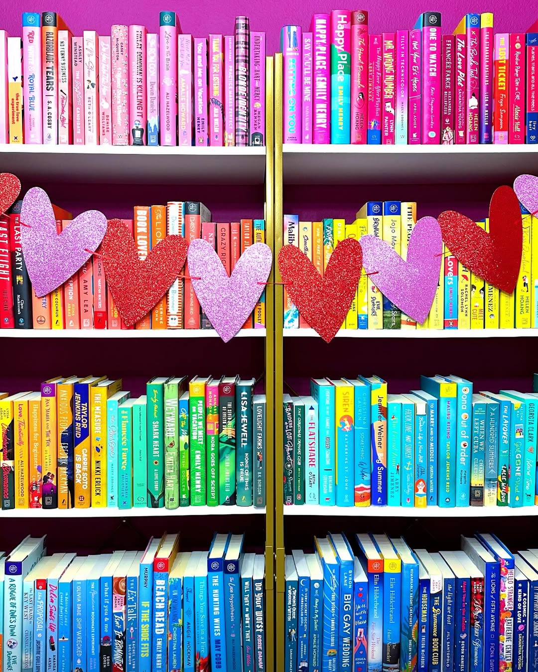 Bookshelves of color-coordinated books arranged in a rainbow gradient with a garland of red, pink, and purple glitter hearts hanging across the middle. The background wall is bright pink.