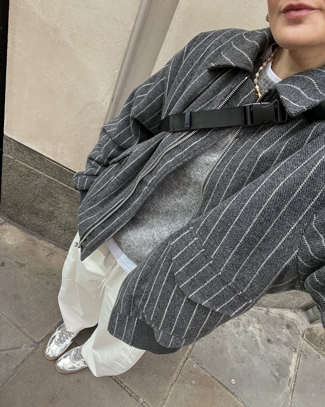 A person wearing a gray pinstripe jacket, gray sweater, white pants, metallic silver shoes, and a black crossbody strap stands on a city sidewalk—capturing one of those awkward moments when their face is partially out of frame.