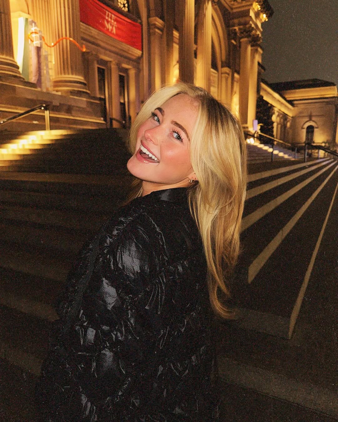 A smiling blonde woman in a black jacket stands at night on the steps of a grand, well-lit building with columns. The scene appears lively and elegant.