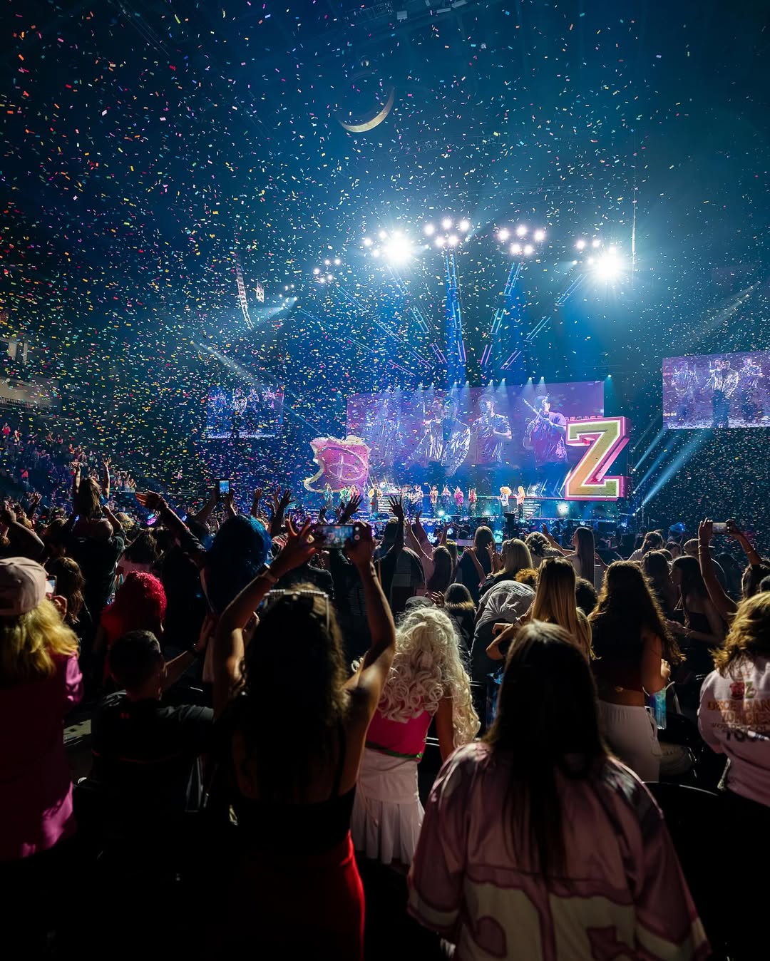 A large crowd enjoys the Worlds Collide concert special with confetti falling, bright stage lights, and a giant letter Z on stage. People in costumes cheer and raise their arms, creating a lively, festive atmosphere.