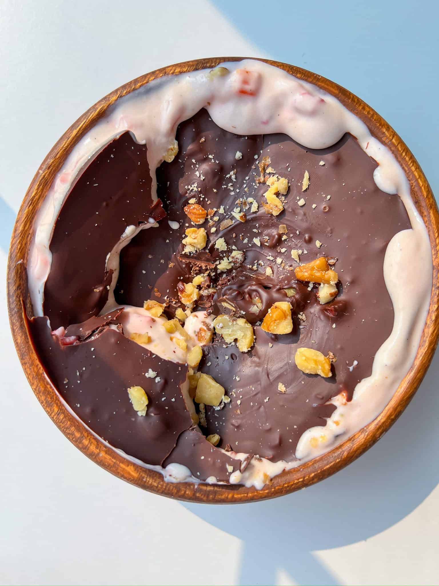 A wooden bowl filled with yogurt topped with a cracked layer of chocolate and sprinkled with chopped nuts, photographed from above on a white surface—perfect inspiration for healthy snack recipes or delicious after school snacks.