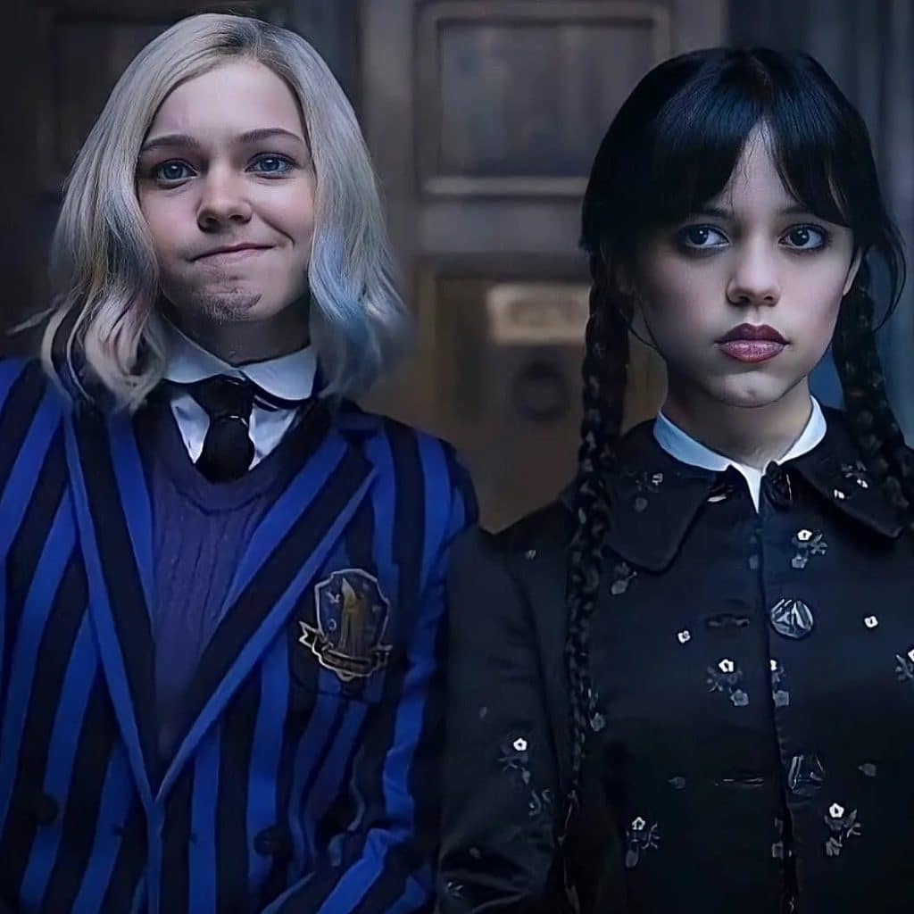 Two young women stand side by side indoors. One has blonde hair, wears a blue striped jacket with a crest, and smiles. The other has long dark braids, wears a black dress with a white collar, and looks serious.