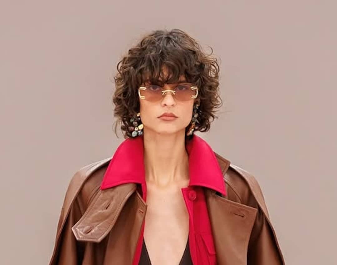 A person with curly dark hair wears sunglasses, bold earrings, and a brown coat with a bright red collar, channeling full-glam vibes against a plain beige background.