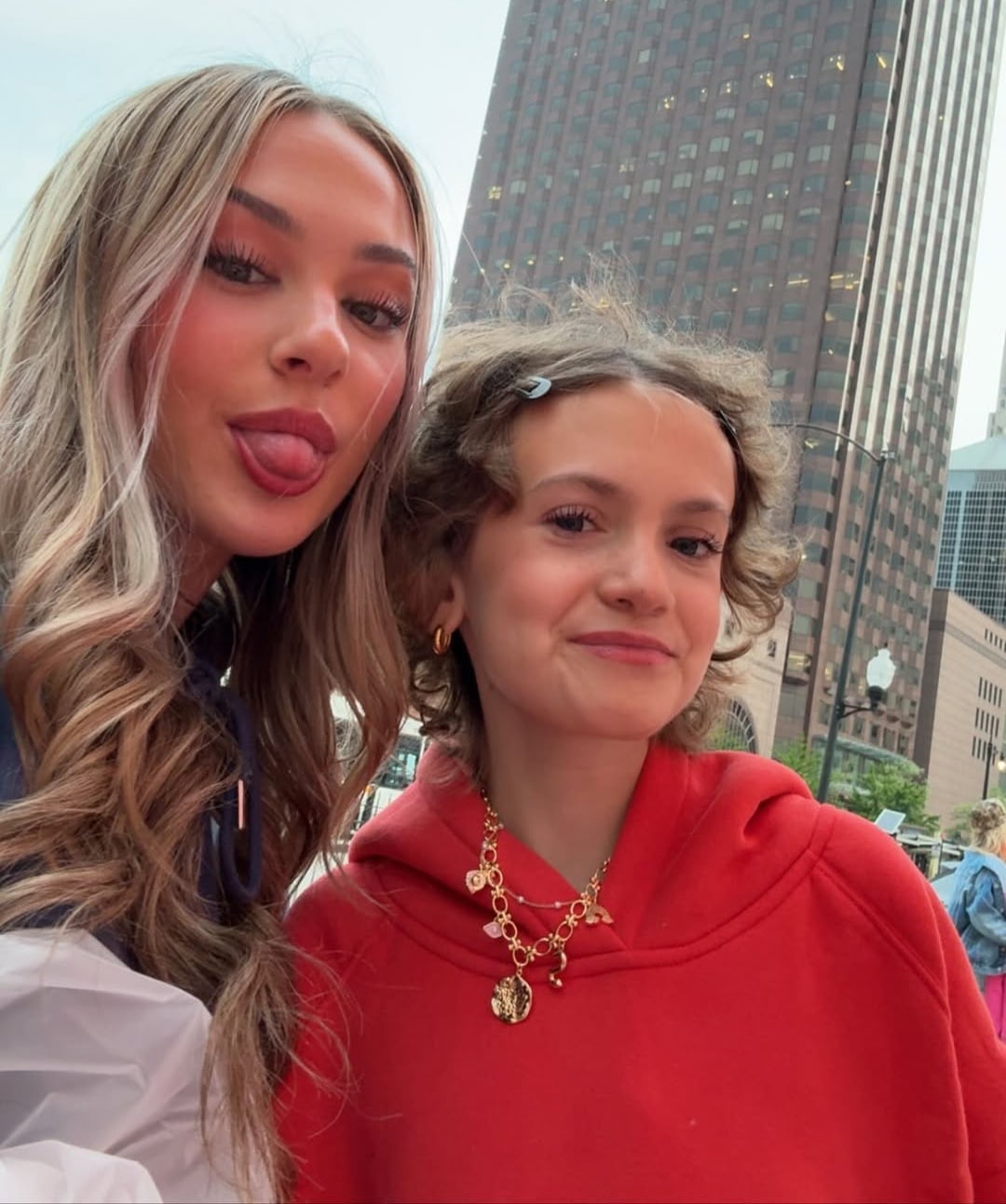 Two young women pose for a selfie outdoors in a city—one, possibly Gianna Joyce, sticks her tongue out while the other in a red hoodie and gold jewelry smiles softly. A tall building rises behind them, perfect for sharing in their group chat.