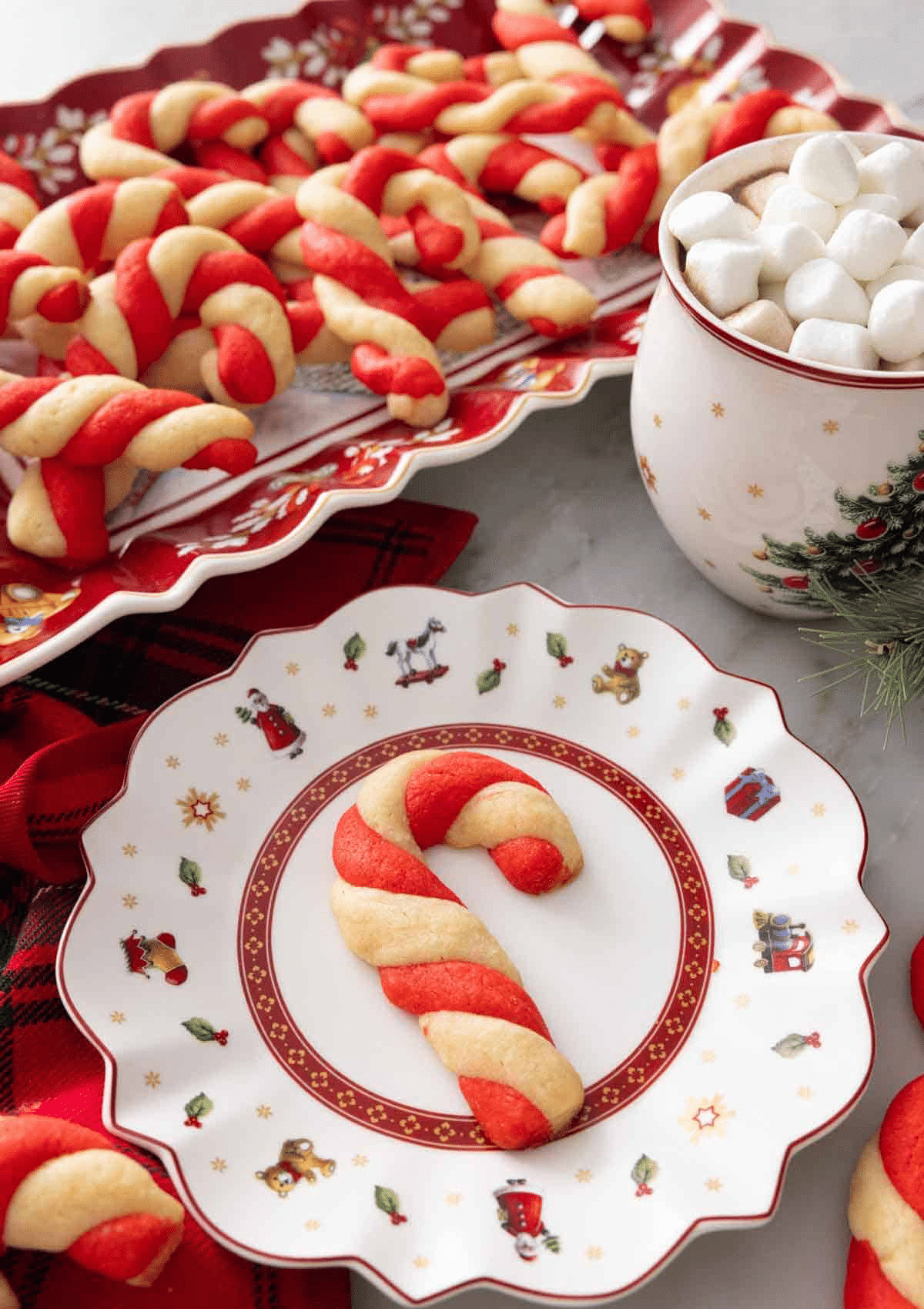 A candy cane-shaped Christmas cookie with red and white swirls sits on a festive plate, surrounded by trays of Christmas cookies and a mug filled with mini marshmallows, all set on a holiday-themed table perfect for holiday baking.