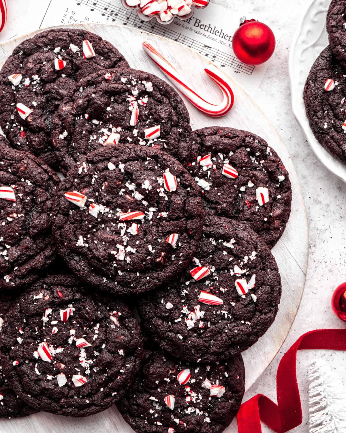 A plate of chocolate cookies topped with crushed peppermint candy and sea salt, surrounded by candy canes, red ornaments, and a red ribbon, evokes a festive holiday atmosphere and inspires dough-lightful Christmas cookie recipes.