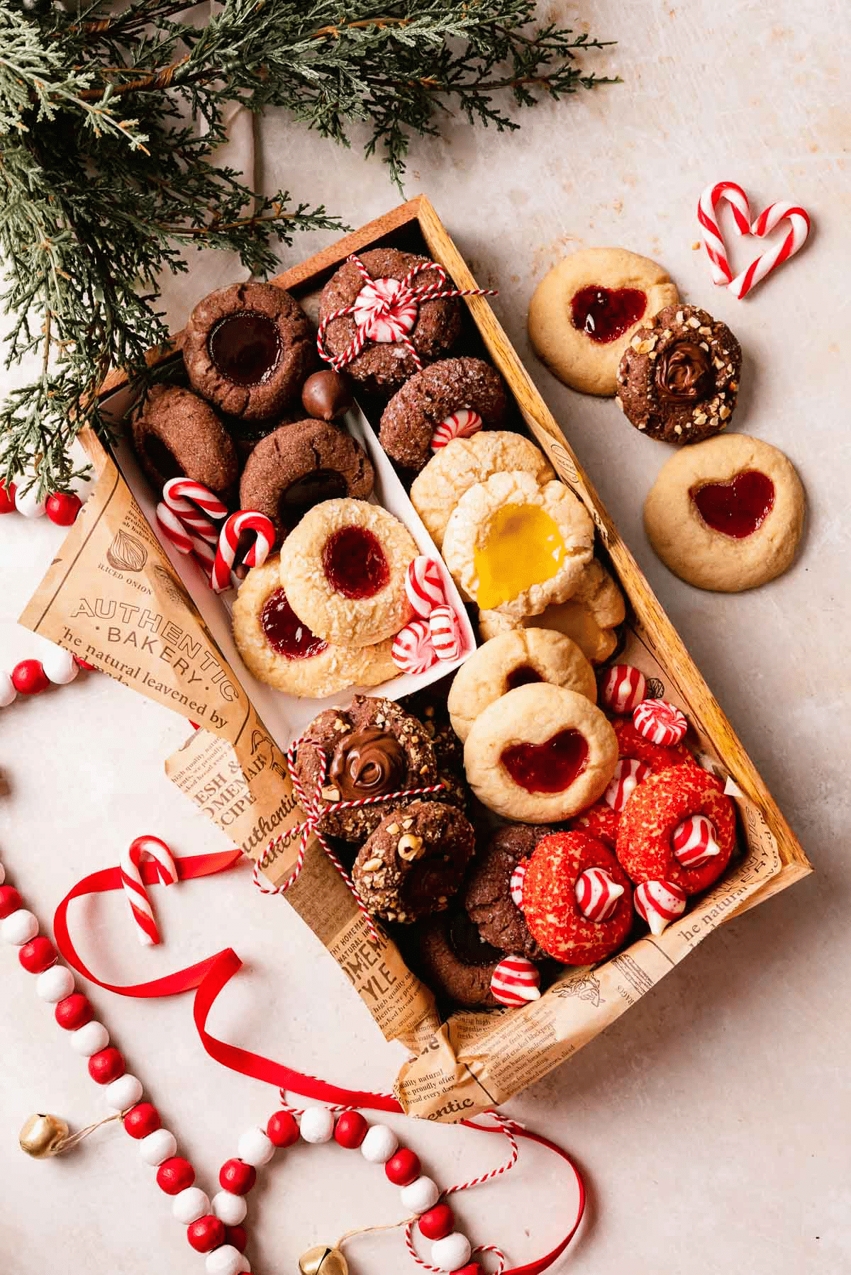 A wooden box filled with assorted Christmas cookies—including thumbprint, chocolate, and sprinkle varieties—is decorated with candy canes and greenery. Extra cookies and festive beads are arranged around the box on a light surface.