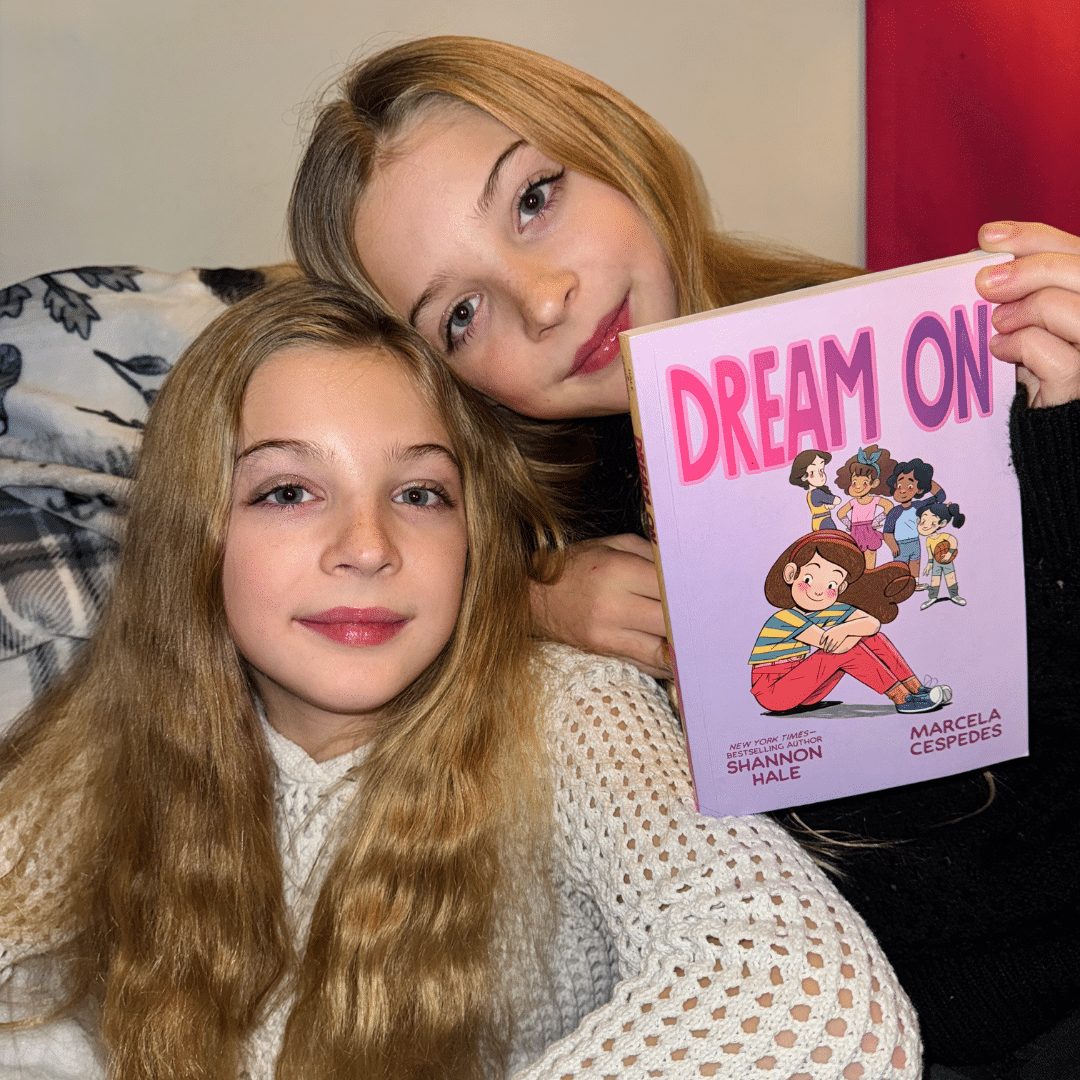 Two young girls with long blonde hair sit closely together. One girl smiles and holds up a colorful book titled “DREAM ON.” Both appear happy and cozy in a warmly-lit room.