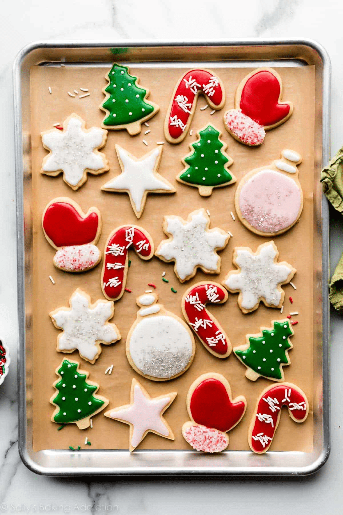 A baking tray lined with parchment paper holds decorated Christmas cookies shaped like stars, snowflakes, trees, candy canes, mittens, and ornaments—perfect for holiday baking—frosted in red, green, white, and pink colors.