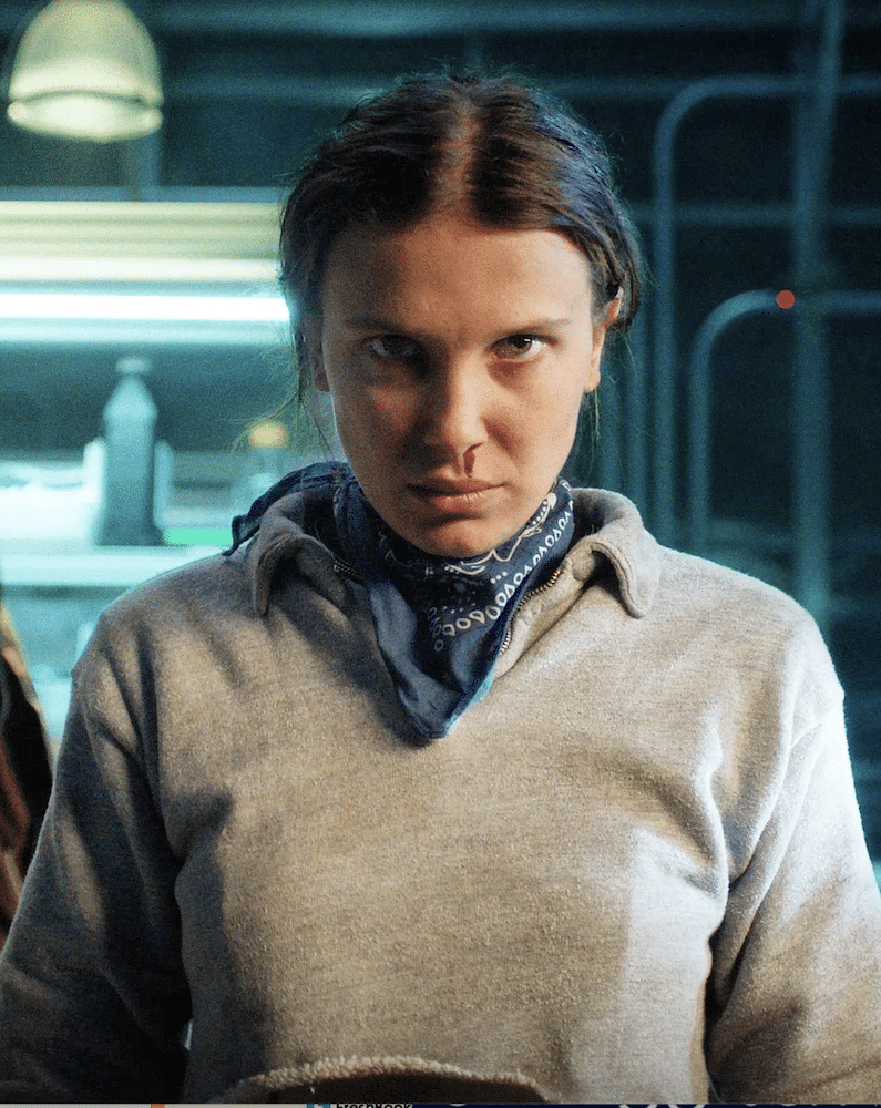 A young girl with a serious expression and a nosebleed stands indoors, resembling a character from Stranger Things, wearing a gray sweatshirt and dark bandana, with blue lighting and blurred objects in the background.