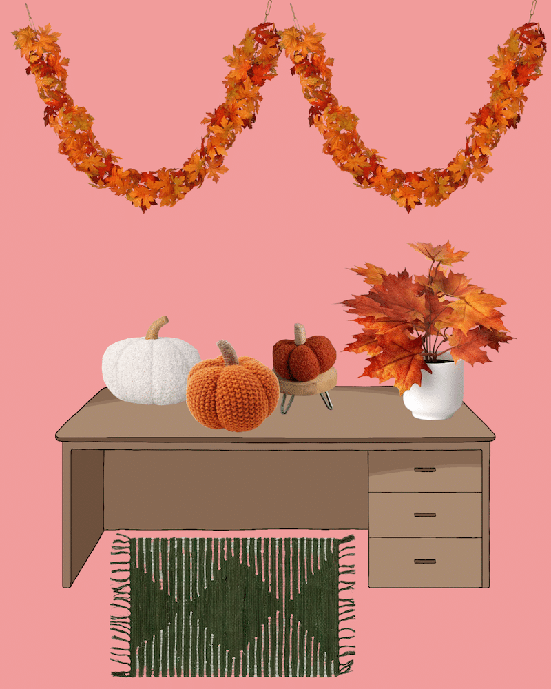 A brown desk with three pumpkins—one white, two knitted—in front of a vase of autumn leaves. An autumn garland hangs above, and a green patterned rug lies beneath, all set against a pink background.