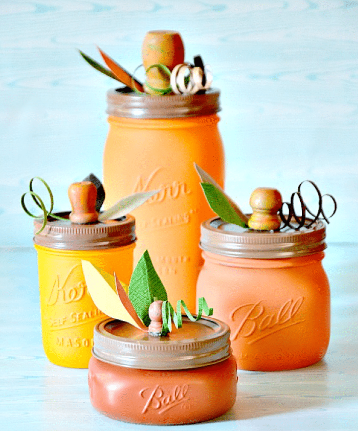 Four mason jars painted in autumn shades of orange are decorated to look like pumpkins, with lids topped by small wooden knobs, green paper leaves, and curly paper stems—perfect for charming seasonal home decor ideas.