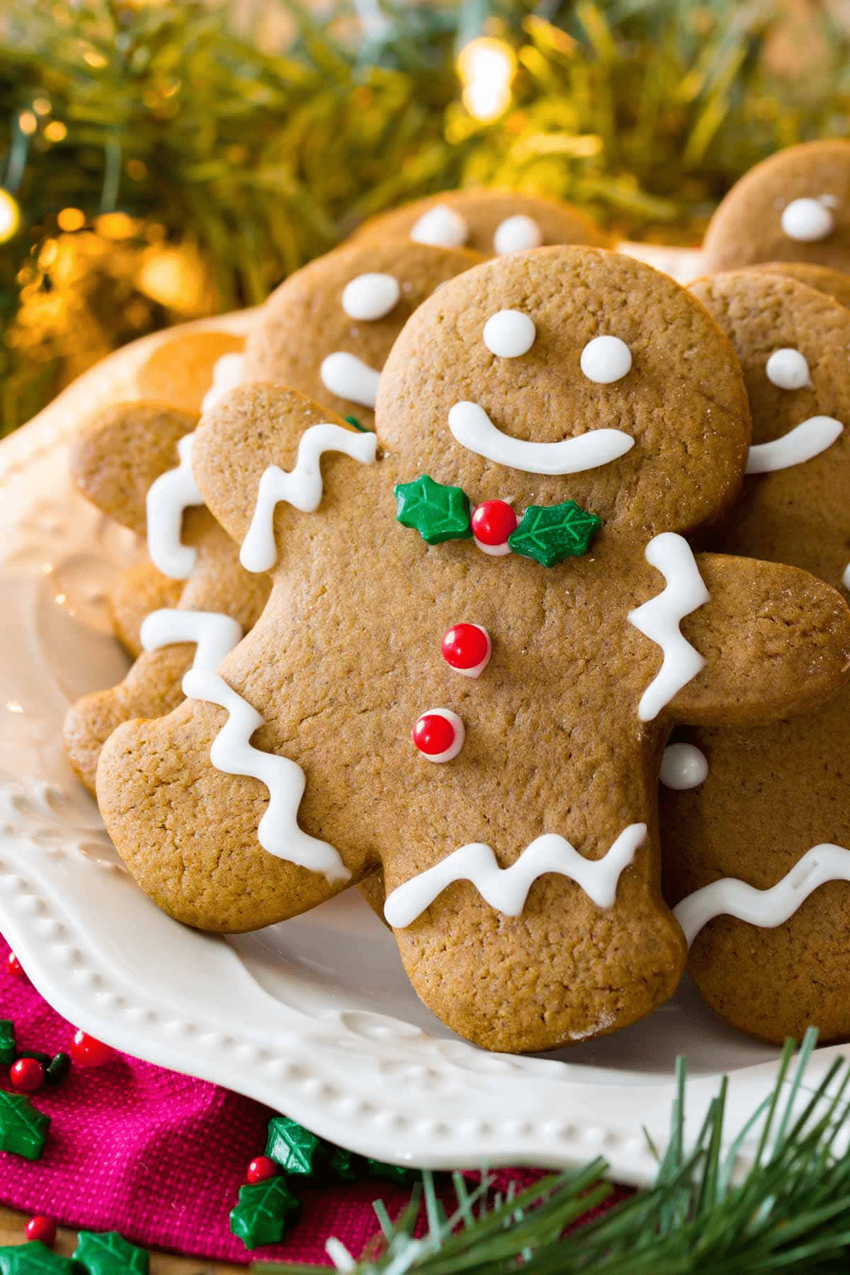 A plate of decorated Christmas cookies shaped like people, with white icing details, red buttons, and green holly decorations, set on a festive table with greenery and holiday lights—perfect inspiration for your holiday baking.