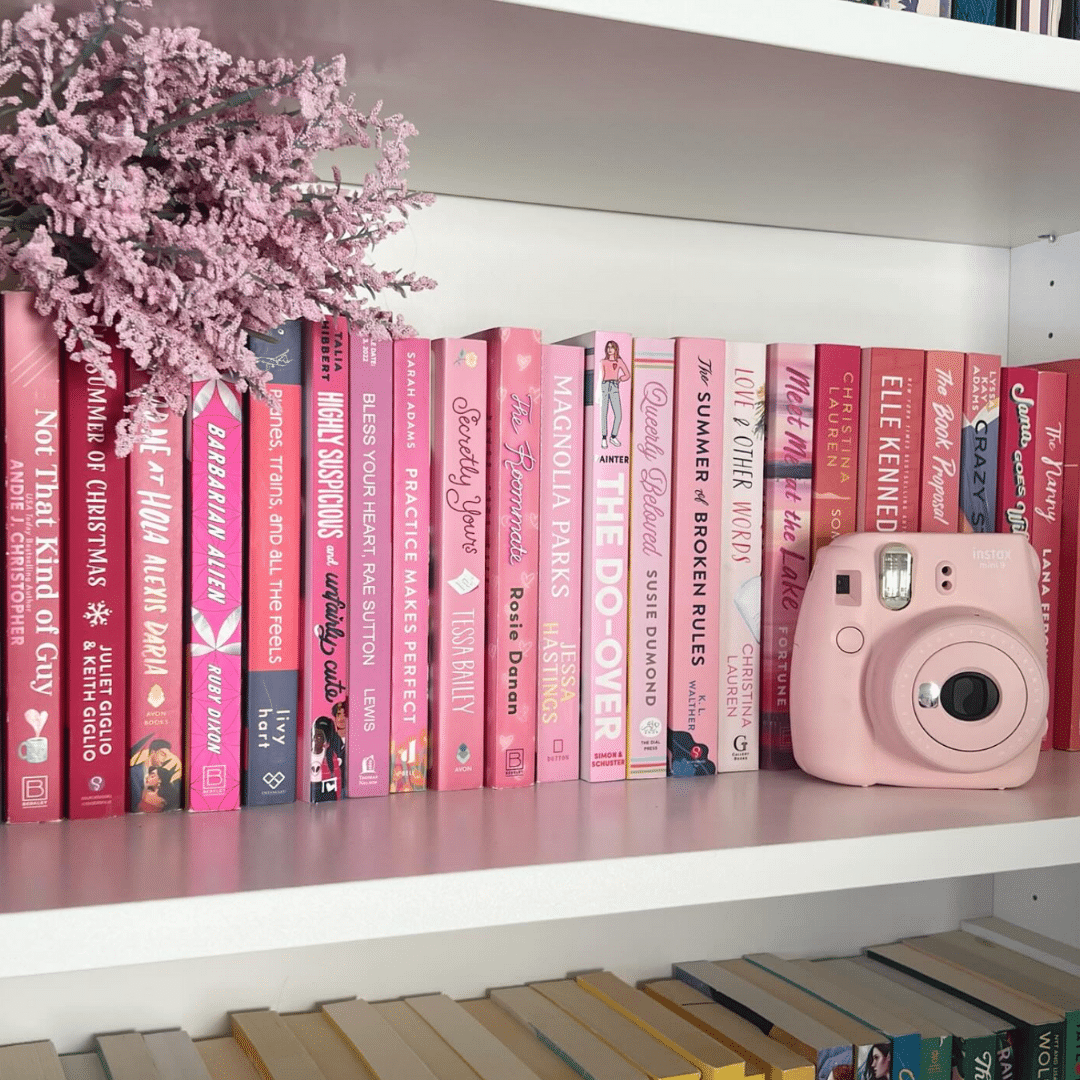 A white bookshelf displaying a row of pink-covered books, a bouquet of pink flowers in a vase, and a pink instant camera, with a shelf of yellow and blue books visible below.